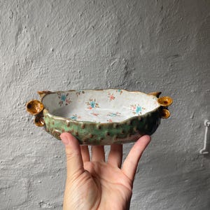 Könnte beinhalten: Eine flache Keramikschale mit einer grünen, strukturierten Außenseite, die kleine braune Erhebungen und dekorative braune pilzartige Formen an den Seiten aufweist. Das Innere ist weiß mit einem Muster aus kleinen blauen und orangen Blumen.