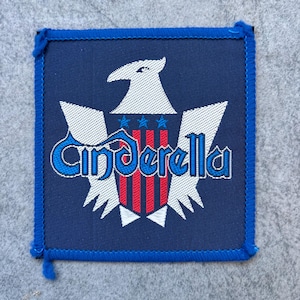Pode incluir: Um emblema bordado quadrado com fundo azul escuro e borda azul costurada. O design apresenta uma águia branca com três estrelas azuis sobre um escudo com listras vermelhas e brancas. A palavra Cinderella está escrita em letras azuis.