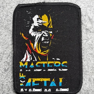 May include: A rectangular black fabric patch with a stitched border featuring a graphic of a stylized face in yellow, white, and red. Below the face, the text reads MASTERS OF METAL in yellow and blue lettering with white accents.