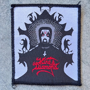 May include: A rectangular woven patch with a black border featuring a white background and a black graphic illustration of a figure with face paint. The design includes red text reading King Diamond, a small white inverted cross, and two small figures.