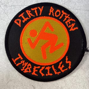 Op de afbeelding: Een ronde geborduurde patch met een zwarte rand en achtergrond. Het heeft een centrale gouden cirkel met een oranje rennend stokfiguurtje. De tekst "DIRTY ROTTEN IMBECILES" is in oranje letters rondom het midden geborduurd.