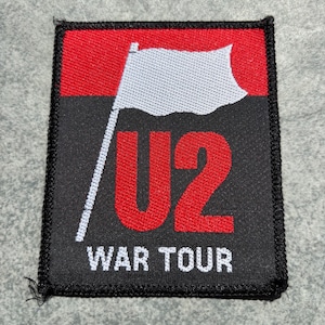 Pode incluir: Um emblema bordado retangular apresentando uma bandeira branca em um mastro contra um fundo vermelho e preto. O texto U2 é exibido em letras vermelhas grandes, com o texto branco WAR TOUR posicionado abaixo na seção preta do emblema.