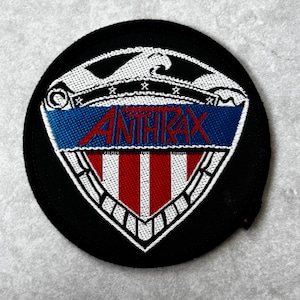May include: A circular embroidered patch featuring a shield design with a black background. The shield displays a white eagle at the top, a blue horizontal band with the red text ANTHRAX, and a lower section with vertical red and white stripes.