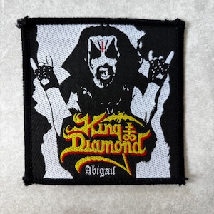 May include: A square embroidered patch featuring a black and white graphic of a figure with face paint and a red cross symbol on the forehead. The text King Diamond is in yellow and red, with the word Abigail in white below the figure.