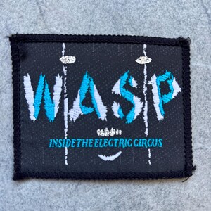Puede incluir: Un parche rectangular negro bordado que presenta el texto W.A.S.P. en una fuente desgastada de color blanco y azul. Debajo del nombre de la banda, el texto INSIDE THE ELECTRIC CIRCUS está bordado en azul. El diseño incluye líneas verticales blancas y un pequeño elemento gráfico blanco.