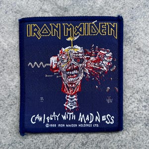 May include: A square embroidered patch with a dark blue background featuring the yellow text IRON MAIDEN at the top. The center shows a graphic of a skull-like figure with red and white details. The white text CAN I PLAY WITH MADNESS is below.