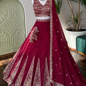 Könnte beinhalten: Ein bordeauxfarbener Lehenga, ein traditionelles indisches Kleidungsstück, das auf einer Schaufensterpuppe präsentiert wird. Das Ensemble verfügt über aufwendige goldene Stickereien auf Rock und Oberteil sowie einen passenden Dupatta. Das Oberteil hat einen V-Ausschnitt und eine dekorative Bordüre. Der Rock hat ein ausgestelltes Design.