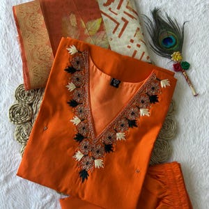 Könnte beinhalten: Ein orangefarbenes, besticktes Kurta-Set mit passendem Dupatta. Das Kurta hat einen V-Ausschnitt mit floralen Stickereien in Schwarz, Weiß und Gold. Eine Pfauenfeder und ein dekorativer Stab sind an der Seite platziert. Das Set wird auf einer weißen Oberfläche präsentiert.