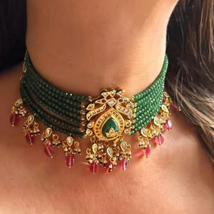 Könnte beinhalten: Eine mehrreihige Halskette mit kleinen grünen Perlen und einem zentralen goldfarbenen Anhänger mit einem tropfenförmigen grünen Stein. Das Schmuckstück ist mit hängenden rosa Tropfenperlen und kleinen weißen Perlen am unteren Rand verziert.