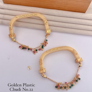 Könnte beinhalten: Zwei Armreifen aus beigem Kunststoff mit goldfarbenen Metallakzenten und hängenden Ketten mit kleinen roten und grünen Perlen. Die Armreifen sind als Golden Plastic Chudi No.22 beschriftet. Jedes Stück hat eine gebogene, halbstarre Struktur.