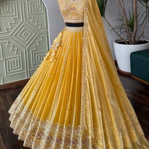 Puede incluir: Un vibrante vestido formal amarillo con intrincados bordados y adornos. El corpiño y la falda presentan patrones detallados, con una falda fluida y un chal a juego. El vestido se exhibe en un maniquí, mostrando su elegante diseño.