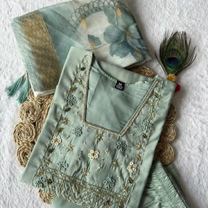 Könnte beinhalten: Ein hellgrünes, besticktes Tunika- und Hosen-Set mit einem Schal mit Blumenmuster. Die Tunika hat aufwendige Blumenstickereien in Weiß, Gold und Blau. Eine Pfauenfeder sorgt für eine dekorative Note.