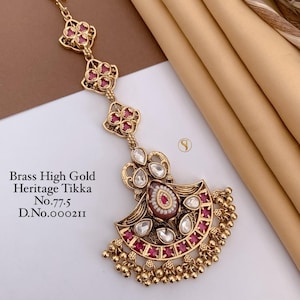 May include: A brass high gold heritage tikka featuring a central fan-shaped pendant with red stones, white accents, and dangling gold beads. The piece includes a chain with three floral-shaped links and a hook. Text reads: Brass High Gold Heritage Tikka No.77.5 D.No.000211.