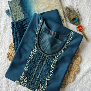 Könnte beinhalten: Türkisfarbenes besticktes Kurta-Anzug-Set. Das Oberteil hat aufwendige Blumenstickereien mit Pailletten und Perlen. Das Set enthält eine passende Hose und einen gemusterten Dupatta. Eine Pfauenfeder und ein dekorativer Gegenstand sind ebenfalls sichtbar.