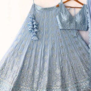 Könnte beinhalten: Hellblaues Lehenga-Choli-Set mit Verzierungen. Das Set besteht aus einem Rock, einem ärmellosen Oberteil mit V-Ausschnitt und einem passenden Dupatta. Rock und Oberteil sind mit aufwendiger silberner Stickerei und Spiegelarbeiten verziert, was einen schimmernden Effekt erzeugt.
