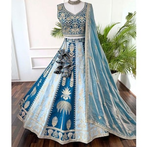 Puede incluir: Un conjunto lehenga choli bordado en azul verdoso y azul claro. La parte superior sin mangas y la falda presentan patrones florales dorados. Una dupatta azul claro a juego con ribete dorado completa el conjunto. El atuendo se exhibe en un maniquí.