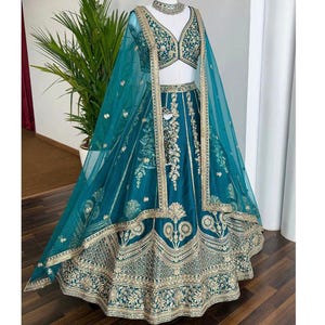 May include: Teal-colored formal gown with intricate gold embroidery. The gown features a matching top, skirt, and sheer shawl. The design includes detailed patterns and embellishments, creating an elegant and sophisticated look. The gown is displayed on a mannequin.