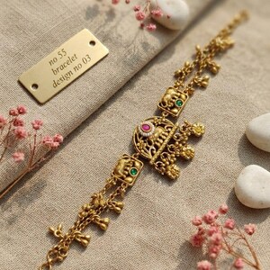 Könnte beinhalten: Ein goldfarbenes Metallarmband mit filigranen Details, kleinen hängenden Anhängern und quadratischen Gliedern mit Elefantenmotiven. Das Mittelstück ist mit einem rosa Stein und weißen Perlen verziert, während grüne Steine die Elefantenglieder akzentuieren.