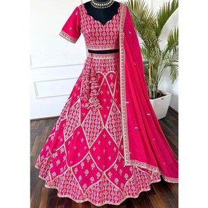 May include: A vibrant fuchsia-colored formal dress with intricate gold embroidery. The set includes a short-sleeved top, a long skirt, and a matching scarf. The design features a detailed pattern and a scalloped hemline, perfect for special occasions.