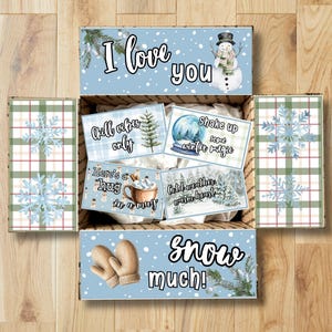 May include: A cardboard gift box with light blue flaps featuring white snowflakes, a snowman, and mittens. The text reads I love you snow much. Inside are four rectangular cards with winter graphics and phrases like Chill vibes only and Here's a hug in a mug.