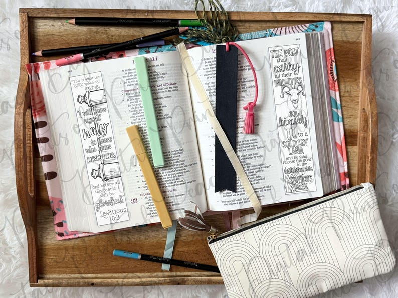 May include: An open book with two illustrated bookmarks featuring text from Leviticus, including a goat drawing and scroll imagery. The book is surrounded by colored pencils, a black bookmark with a pink tassel, and a white pencil case with an arch pattern.