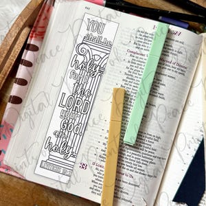 May include: A rectangular paper bookmark featuring a black and white line art design of a classical column with the text YOU shall be holy for I the LORD your God am holy LEVITICUS 19:2. The bookmark rests on an open book with two plastic markers.