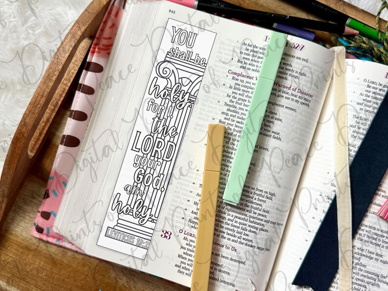May include: A rectangular paper bookmark featuring a black and white line art design of a classical column with the text YOU shall be holy for I the LORD your God am holy LEVITICUS 19:2. The bookmark rests on an open book with two plastic markers.