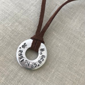 May include: A silver metal necklace with a round pendant featuring the coordinates "N 35.2851° W 063.639°" engraved on it. The necklace is made with a brown suede cord.