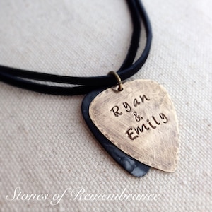 May include: A personalized guitar pick necklace with the names "Ryan & Emily" engraved on a brass-colored pick. The pick is layered over a dark gray pick, suspended from a black cord. The words "Stones of Remembrance" are at the bottom.
