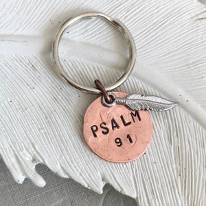 May include: A copper-coloured keychain with a silver key ring and a small silver feather charm. The keychain has the words "PSALM 91" engraved on it.