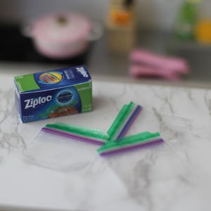 May include: A miniature box of Ziploc sandwich bags with a blue and green design. Three clear plastic sandwich bags with green and purple closures are next to the box. The background is blurred.