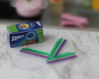 以下が含まれることがあります： 青と緑のデザインの、Ziplocサンドイッチバッグのミニチュアボックス。緑と紫の留め具が付いた3つの透明なプラスチックサンドイッチバッグが箱の隣にあります。背景はぼやけています。