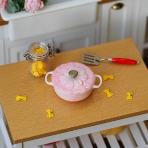 May include: A miniature kitchen scene featuring a pink lidded pot, a small glass jar with yellow contents, and a spatula with a red handle. Several yellow bow-tie pasta shapes are scattered on the wooden surface. The pot has white handles.