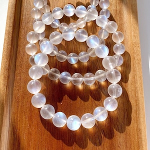 Op de afbeelding: Verschillende heldere, ronde edelsteenarmbanden met een subtiele iriserende glans liggen op een houten dienblad. De kralen zijn aan elkaar geregen, waardoor een delicaat accessoire ontstaat. De armbanden worden verlicht door natuurlijk licht.