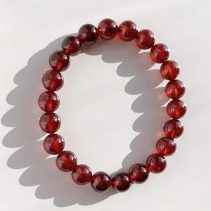 Puede incluir: Una pulsera de cuentas con piedras preciosas redondas y pulidas de color rojo intenso. Las cuentas están ensartadas para formar un círculo completo. Las cuentas, de tamaño uniforme, tienen un acabado brillante. La pulsera se muestra sobre una superficie blanca.