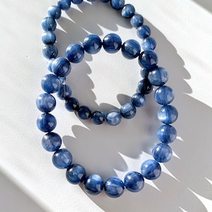 May include: Two blue kyanite beaded bracelets. The bracelets are made of round, polished beads in varying shades of blue. The bracelets are displayed on a white surface with shadows.