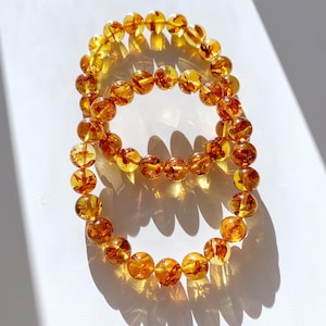 May include: Two amber beaded bracelets are displayed on a white surface. The beads are a warm, translucent orange-yellow color, with visible inclusions. The bracelets are arranged in a circular shape, casting shadows.