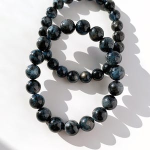 May include: Two beaded bracelets featuring dark blue and black spherical beads. The bracelets are arranged in a figure-eight pattern, with the beads varying in size. The beads have a glossy finish and reflect light, creating a shimmering effect.