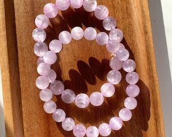 May include: Two light purple beaded bracelets rest on a wooden tray. The bracelets are made of round, translucent beads. The wooden tray has a warm, brown tone, and the bracelets are positioned to catch the sunlight.