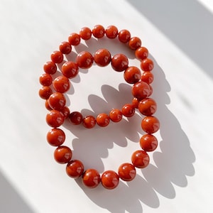 Peut inclure: Deux bracelets de perles rouges sont disposés sur une surface blanche. Les perles sont rondes et ont une finition brillante. Les bracelets sont de tailles différentes, l'un ayant des perles plus grosses que l'autre. L'éclairage crée des ombres.