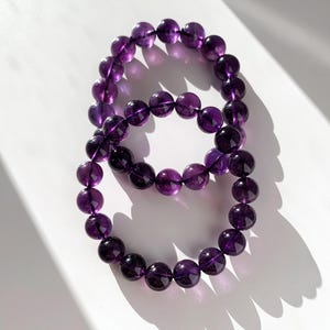Könnte beinhalten: Zwei Armbänder mit Amethystperlen. Die Perlen sind tiefviolett und scheinen glatt und poliert zu sein. Die Armbänder sind in Form einer Acht vor einem weißen Hintergrund angeordnet, wobei Schatten Tiefe verleihen.