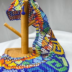 May include: A colorful beaded necklace displayed on a wooden stand. The necklace features intricate patterns in shades of blue, green, yellow, orange, and red. The beads are small and tightly woven, creating a textured surface. The necklace is a vibrant piece of jewelry.