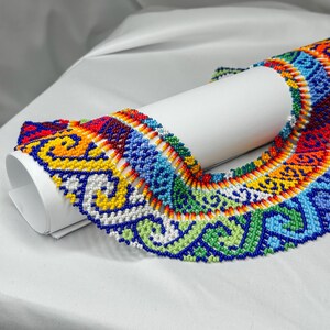 May include: A colorful beaded necklace with intricate patterns in red, yellow, blue, green, and white. The necklace is draped over a white cylindrical object, showcasing its vibrant design and craftsmanship. The beaded jewelry is a striking accessory.