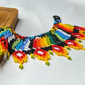 May include: A colorful beaded necklace with a vibrant array of colors including red, orange, yellow, green, blue, and black. The necklace is displayed on a wooden stand, showcasing its intricate design and craftsmanship. The beaded necklace is a statement piece.