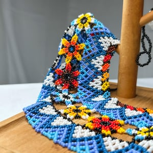 May include: A beaded necklace with a floral design. The necklace features a blue background with white, yellow, orange, and red flower accents. The necklace is displayed on a wooden jewelry stand.