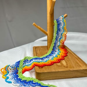 May include: A colorful beaded necklace displayed on a wooden jewelry stand. The necklace features a vibrant rainbow of colors, including red, orange, yellow, green, blue, and white. The beads are small and intricately woven together, creating a textured, wavy pattern.