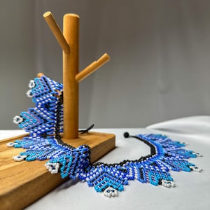 May include: A beaded necklace with a striking blue and white pattern, displayed on a wooden jewelry stand. The necklace features a black base and intricate beadwork, creating a bold, eye-catching design. The stand is made of light-colored wood.