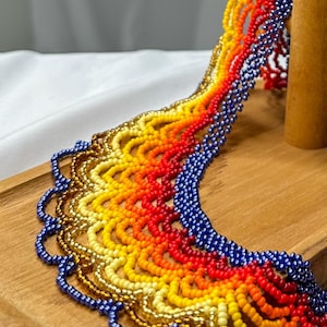 May include: A colorful beaded necklace with a gradient of colors, including gold, yellow, orange, red, and blue. The necklace is displayed on a wooden surface, showcasing its intricate design and craftsmanship. The beads are small and tightly strung.