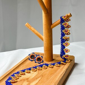 May include: A beaded jewelry set featuring a necklace, earrings, and a bracelet. The jewelry is primarily blue with yellow and red accents. The set is displayed on a wooden tray and a wooden jewelry stand.