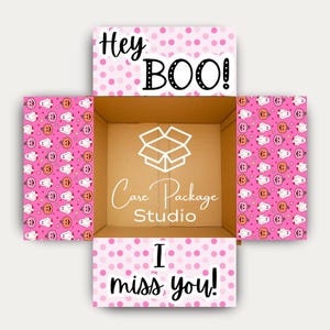 May include: An open cardboard shipping box with interior flaps featuring a pink polka dot pattern, white ghost and orange pumpkin icons, and the text Hey BOO! and I miss you! The center base displays a white box logo and Care Package Studio text.